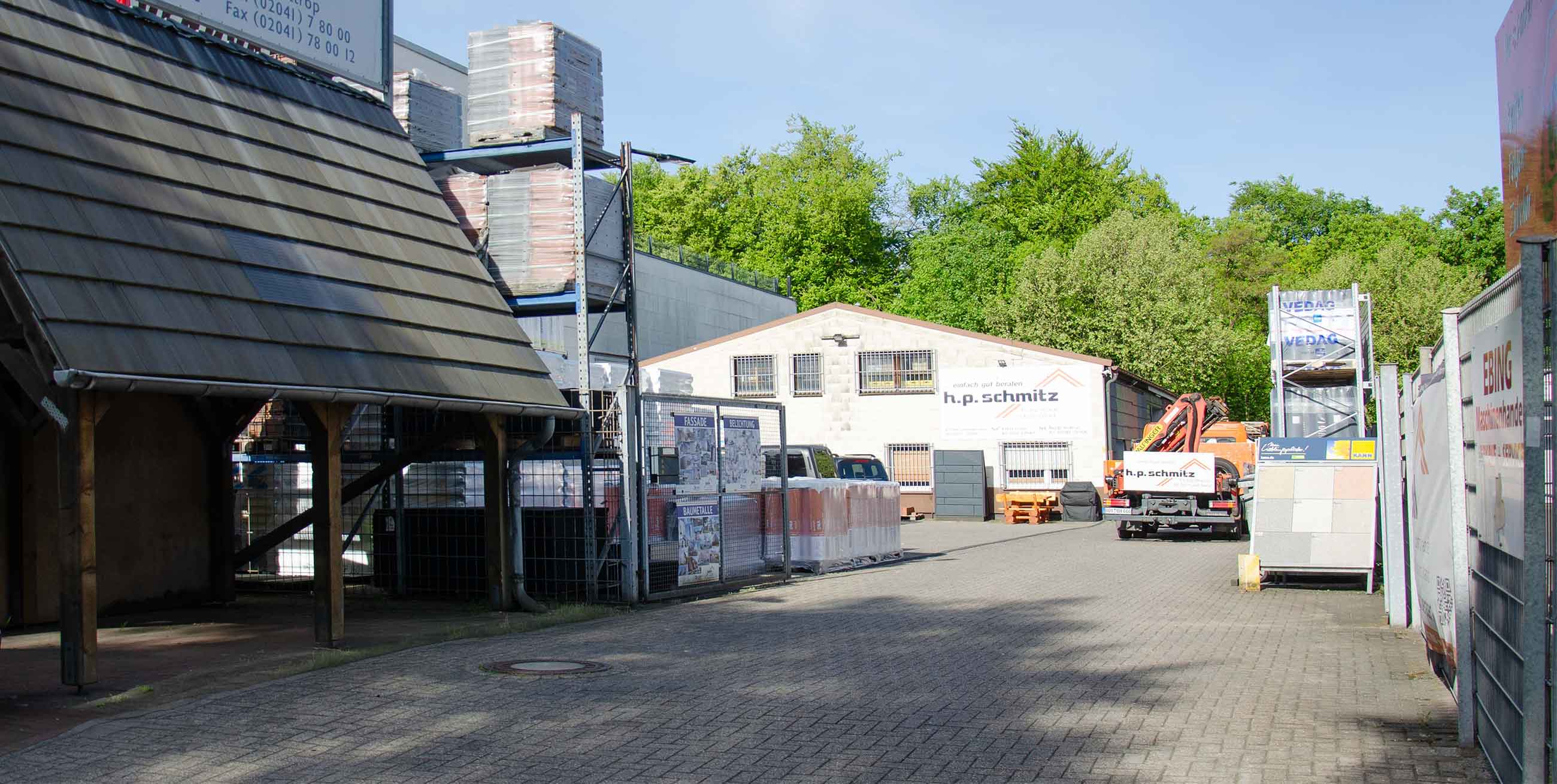 Außenbereich eines Baustoffhändlers mit Lagerhalle, Fahrzeugen und Baustoffen unter blauem Himmel.