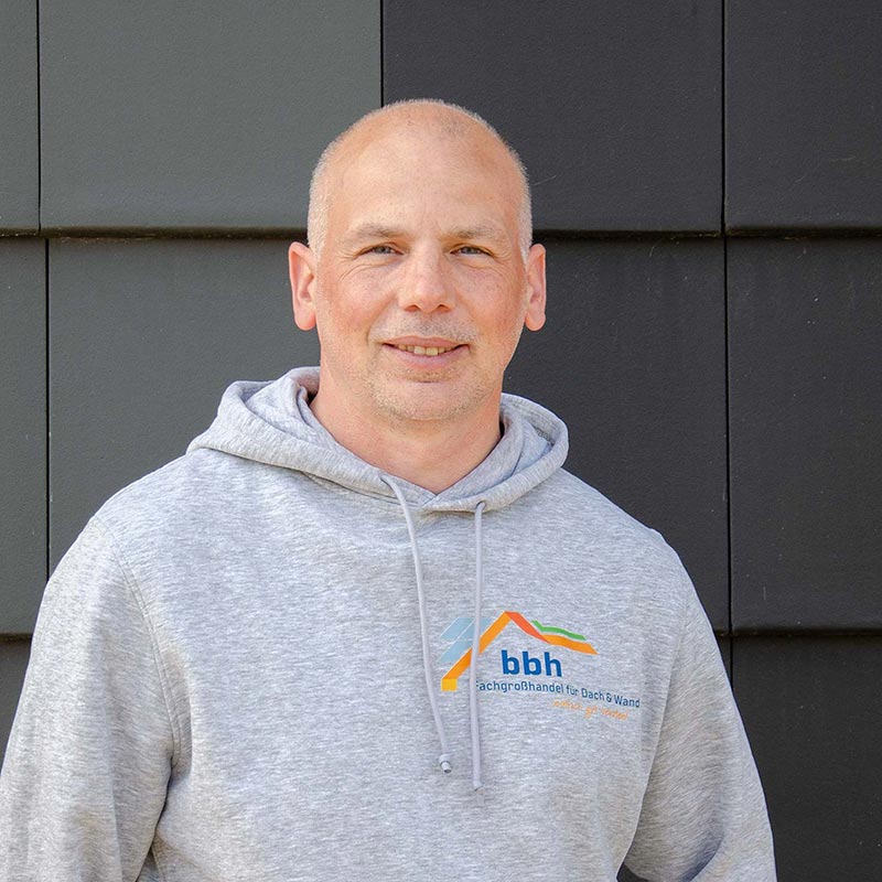 Maik Sypitzki Mann in grauem Kapuzenpullover mit bbh-Logo lächelt vor schwarzer Wand.