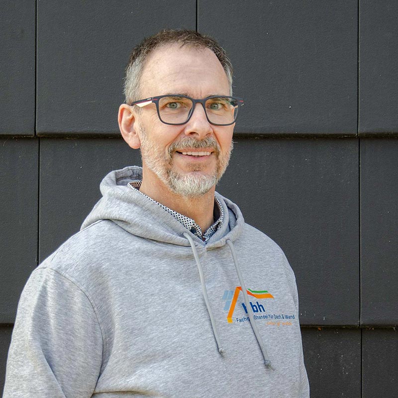 Norbert Buttler Mann mit grauem Kapuzenpullover und Brille lächelt vor schwarzer Wand.
