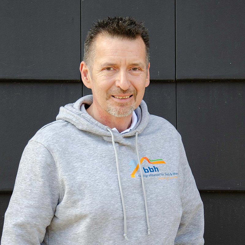 Thomas Nowodworski Mann mit grauem Hoodie vor schwarzer Wand, Hoodie hat bbh-Logo für Dach- und Wasserservice.