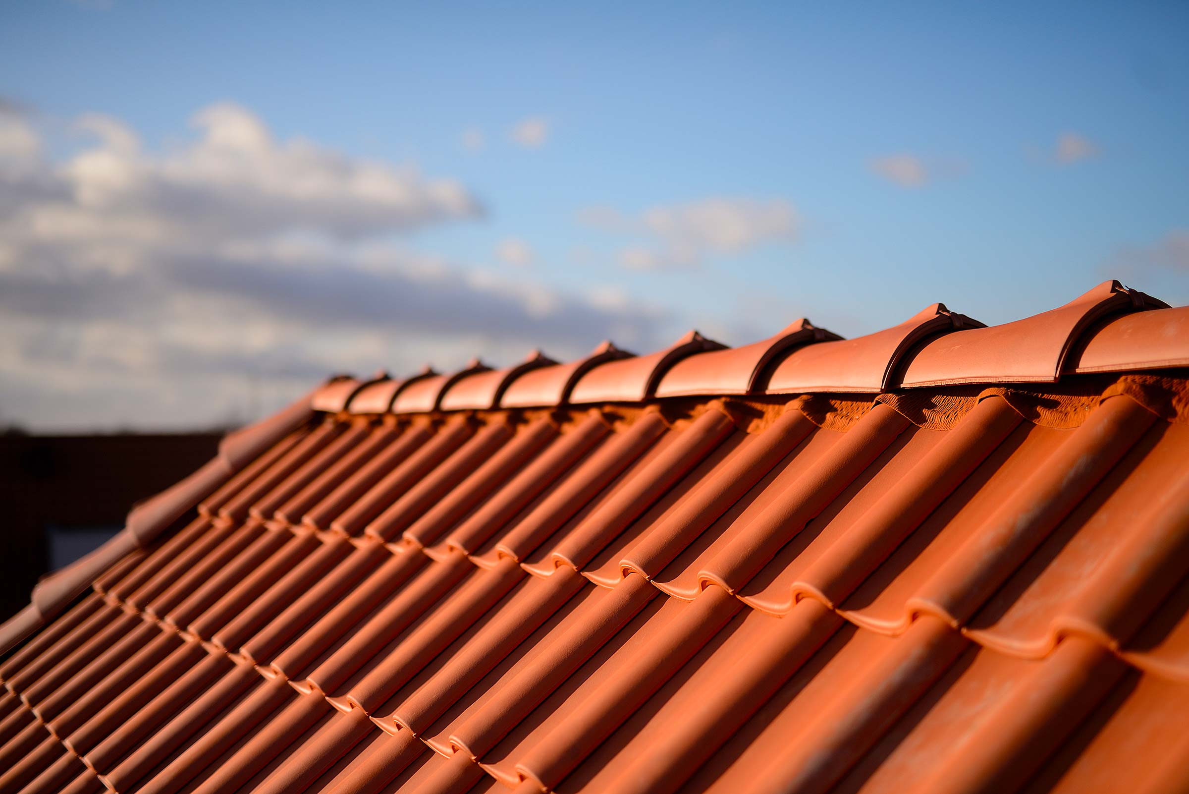Hausdach mit roten Dachpfannen Nahaufnahme eines Dachs mit orangen Ziegeln unter klarem Himmel und vereinzelten Wolken.