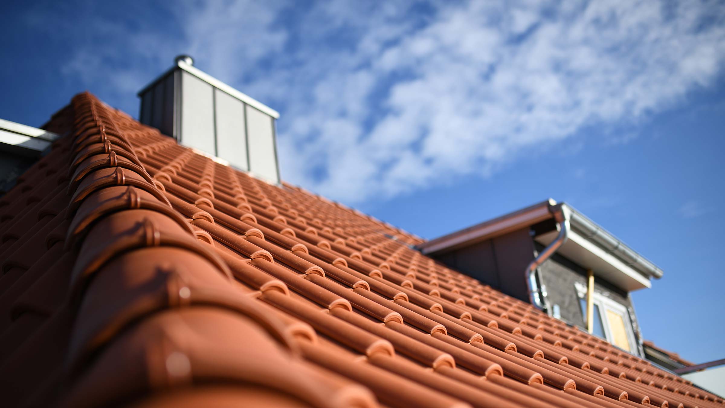 Steildachlösungen mit System Schrägdach mit roten Ziegeln, Dachgaube, Metallrohr und blauem Himmel mit Wolken.