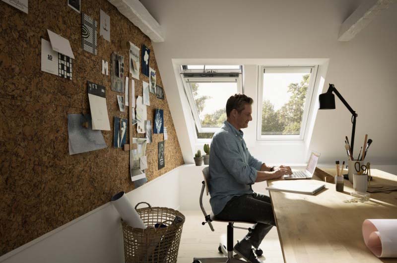 Belichtung & Dachfenster für mehr Wohnqualität Mann sitzt an einem hellen Schreibtisch unter Dachfenstern und arbeitet an einem Laptop.